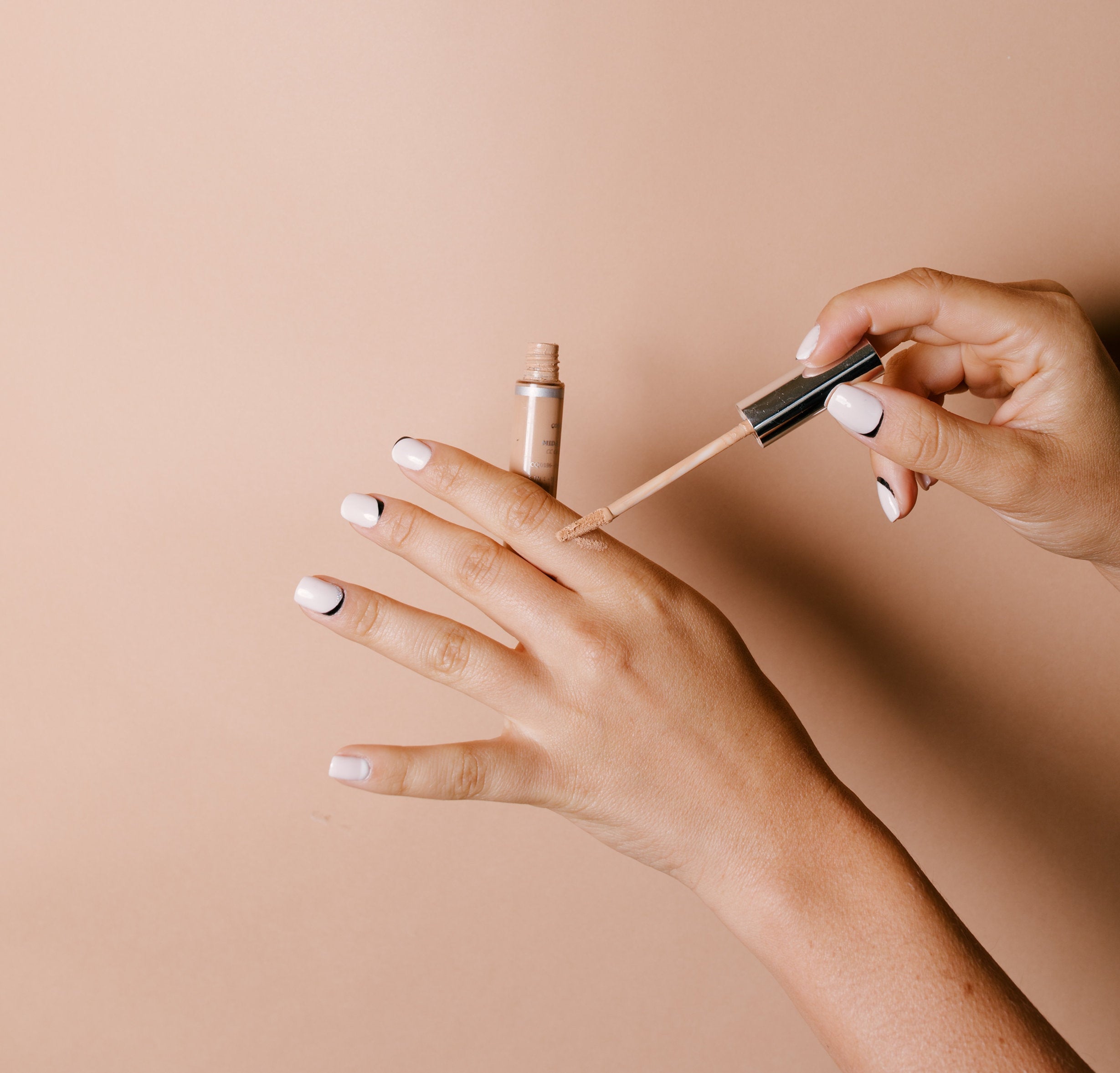 Studio photograph of hands testing liquid makeup from a small bottle with an applicator wand on the skin, displaying white nails with black crescent designs against a plain beige background