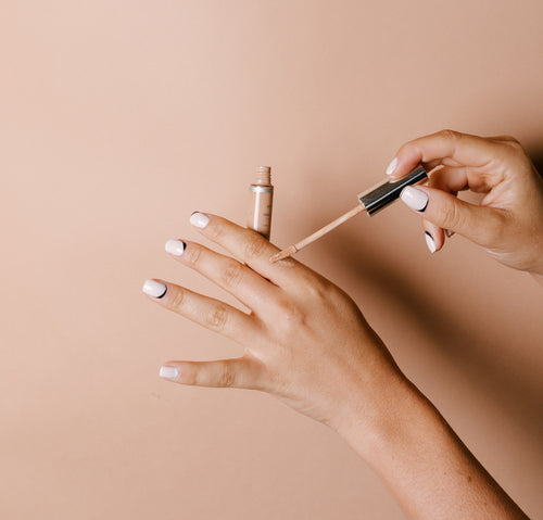 Studio photograph of hands testing liquid makeup from a small bottle with an applicator wand on the skin, displaying white nails with black crescent designs against a plain beige background