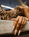 Photography of a woman's hand with long nude coffin nails and two delicate silver rings, resting on a textured concrete surface with a blurred leopard print pattern behind